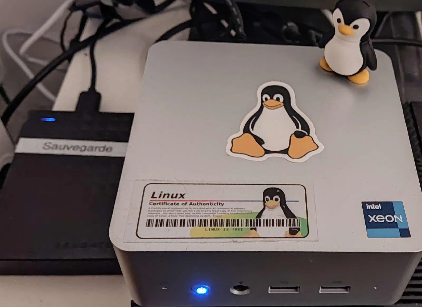 Mini PC prateado com adesivo do Tux, usado como servidor doméstico, ao lado de um HD externo